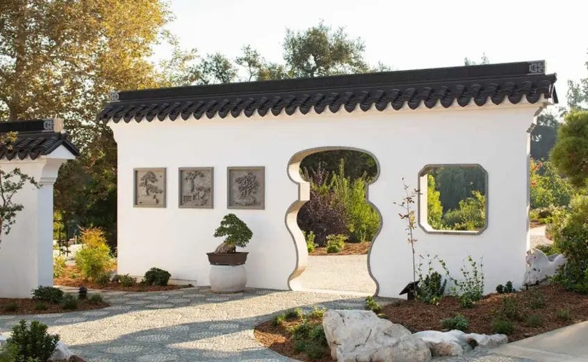 White freestanding wall with a cutout window and door. Three square stone artworks hang on the wall. In front of the wall is a penjing.