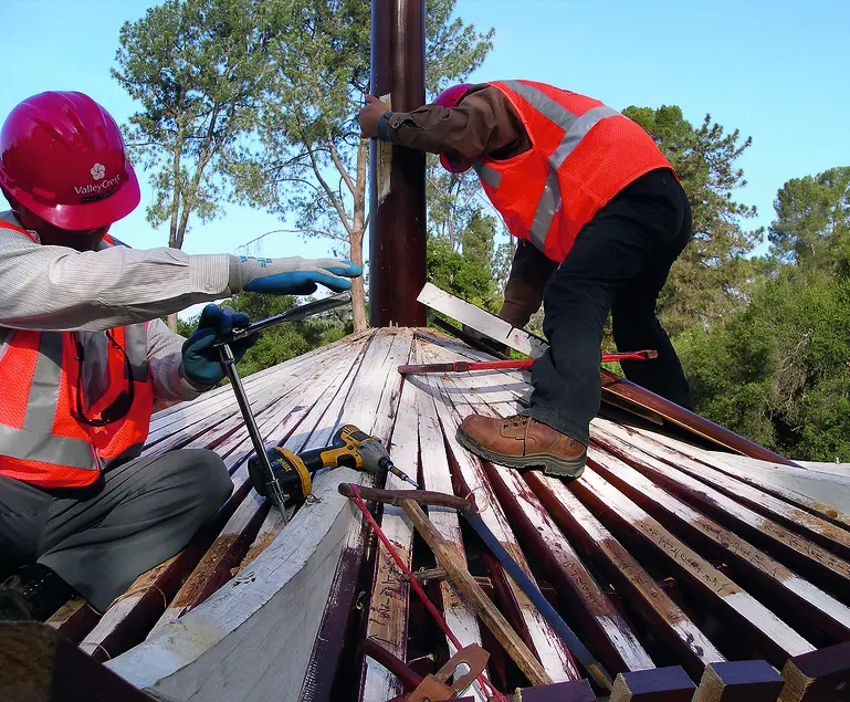 Two people in orange vests and red hard hats work on a partially-finished roof. The roof has wooden slabs.