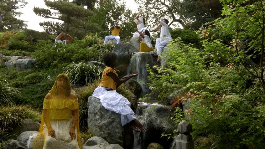 Black and brown people wearing yellow and white sit on rocks at varying levels of a waterfall. Some of the people have their arms outstretched while others have their hands close to their faces. One person in the foreground has their arms to their sides. They are covered in yellow netting and are looking into the camera.