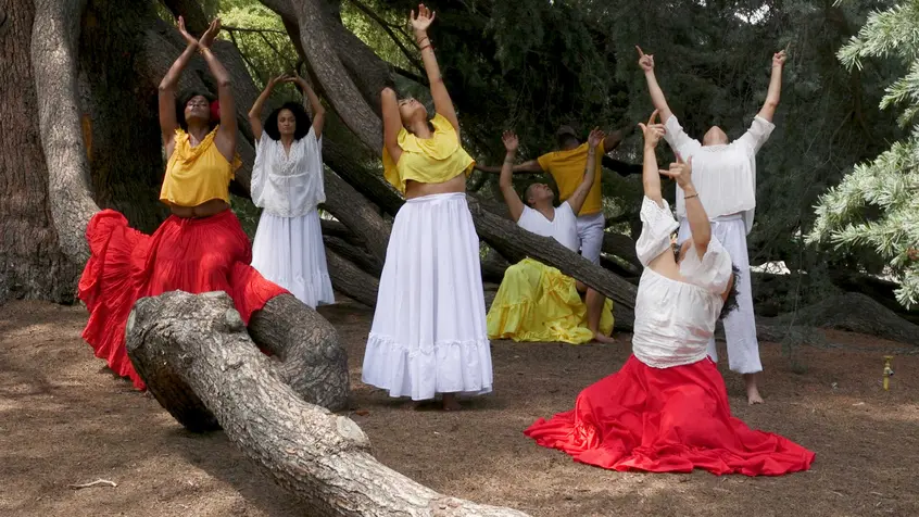 Black and brown people wearing red, yellow, and white sit and stand on the ground with their arms raised above their heads.