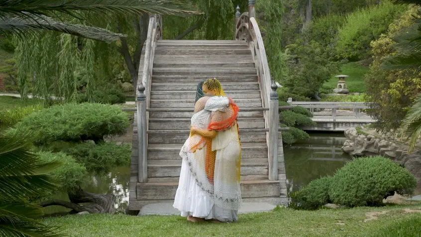 Two people wearing white hold each other in an embrace in front of a domed bridge. Their bodies are covered in an orange and yellow netting. 