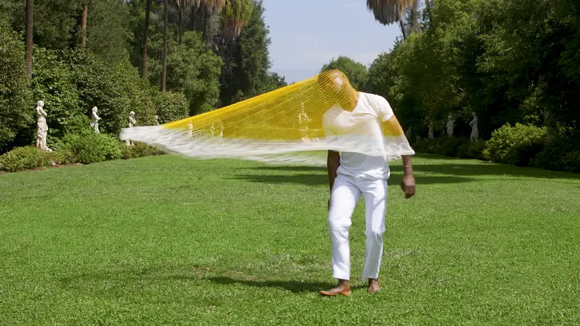 A Black person in white clothing spins a yellow net on their head and shoulders. The person is barefoot on a long, statue-lined lawn.