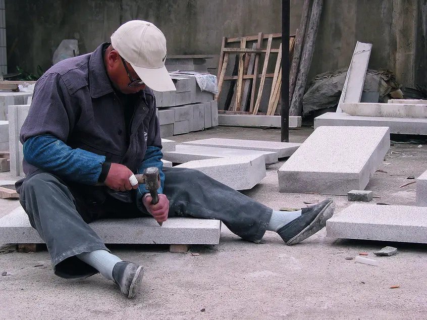 Person sitting on the ground surrounded by rectangular construction materials. The person uses hand tools to work on one of the materials.
