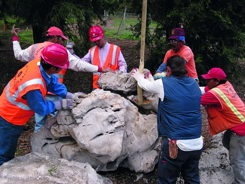Six people, five of whom wear orange vests and hard hats, stand in a circle around a pile of rocks.