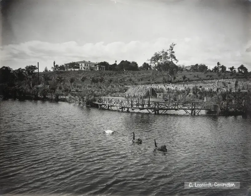 Black and white photograph of a body of water with waterfowl on it. Behind the water is an expanse of land leading up to a mansion on a hill.