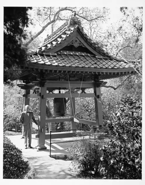 A black and white photograph of a middle aged white man wearing a suit and bow tie rests his left hand on the side of a pagoda-esque bell house.