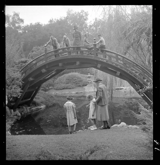 A black and white photograph of a domed wooden bridge in a garden. Four children and one adult stand at the top of the bridge and two children and one adult stand in front of the bridge.
