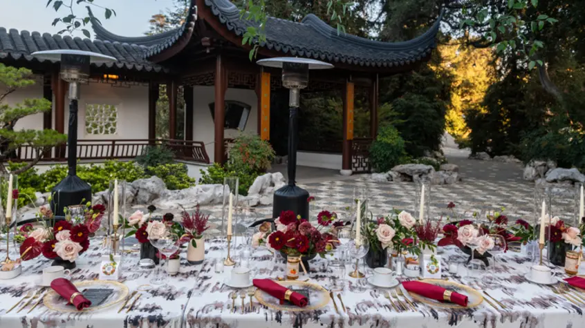 A dining table with place settings of red and light pink set up in an outdoor courtyard.