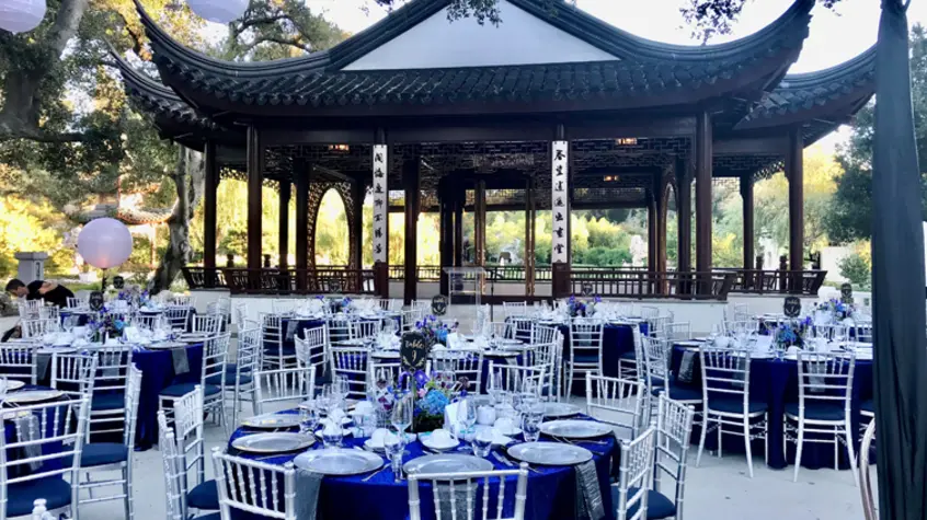 Large, round tables covered in vibrant-blue tablecloths and pewter dinnerware sits in a courtyard in front of a large open pavilion.