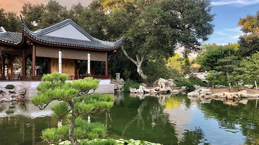A Chinese pavilion stands majestically behind a small lake with a cloudy sky of whites, blues, and amber.