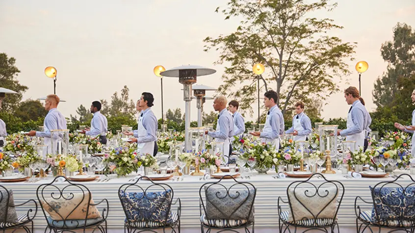 A lineup of wait staff in powder blue shirts and white aprons hold appetizers and plates.
