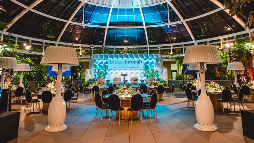 A blue-lit stage with tables, chairs, and oversized floor lamps bring life to an evening event under a glass dome.
