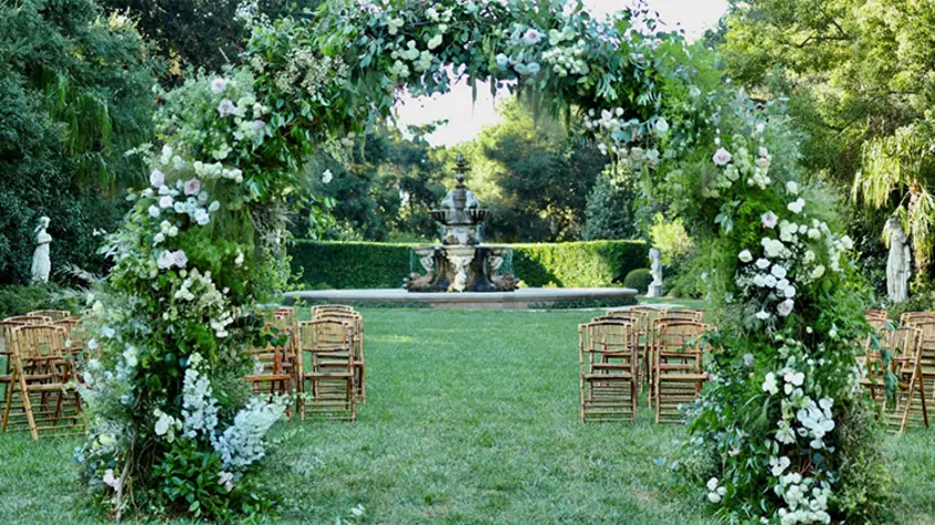 A plant-filled trellis opens up to wooden chairs set up on a green lawn with a large garden fountain in the background.