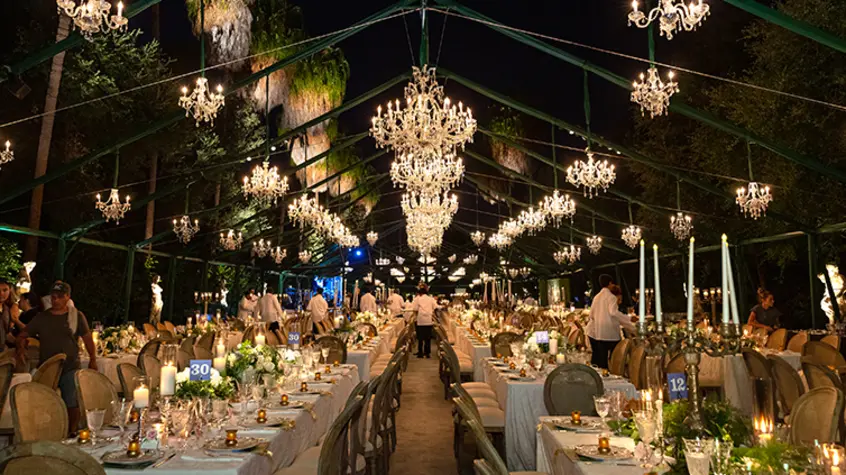 Dozens of small and medium-sized chandeliers hang down from an outdoor pergola brightening up a nightime dinner party setup.