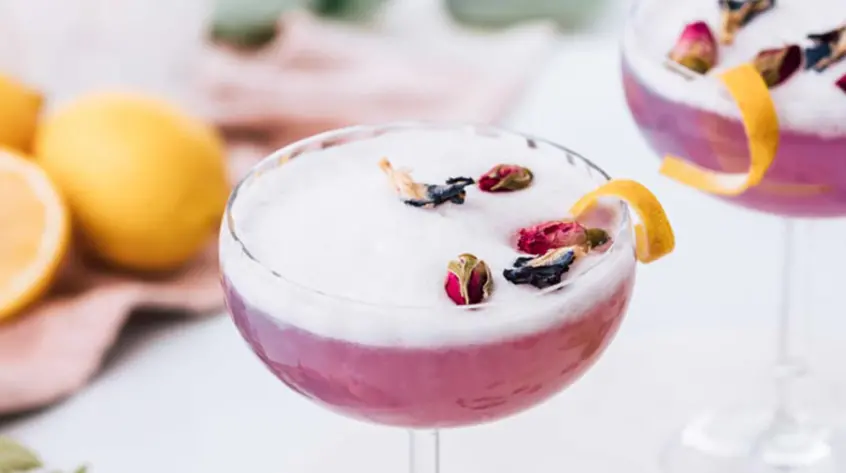 A magenta-colored gin sour sits in fancy stemmed glassware topped with fresh fruit and a lime twist.