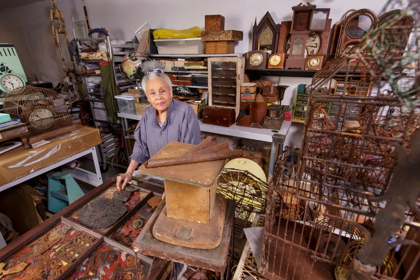 Artist Betye Saar, stands behind a table in her studio, surrounded by various objects.