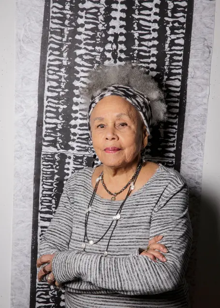 Artist Betye Saar, stands facing the camera with arms crossed in front of a pattern of bodies in silhouette.