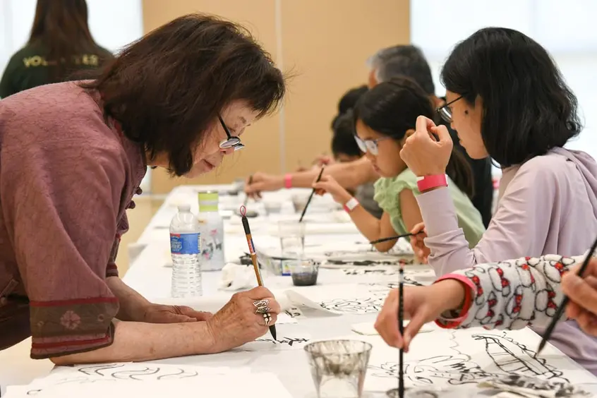 Class instructor paints Chinese characters on a paper in front of students.