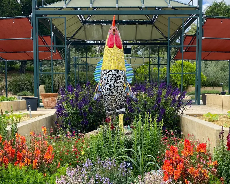 A large sculpture of a rooster, covered in mosaic tiles, surrounded by flowers.