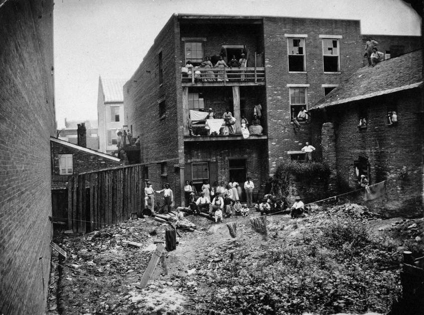 A black and white image of a hospital. Balconies filled with people overlook an open lot.