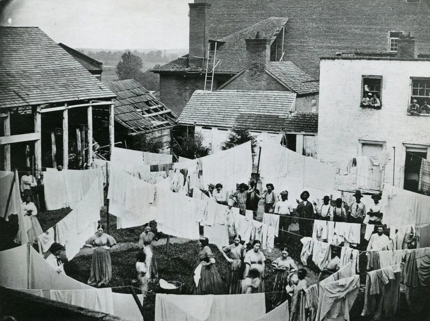 An overhead view of a laundry yard filled with people and hanging fabric, surrounded by buildings.