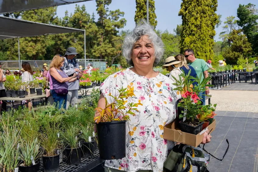 A smiling person holds potted plants near a table with plants.