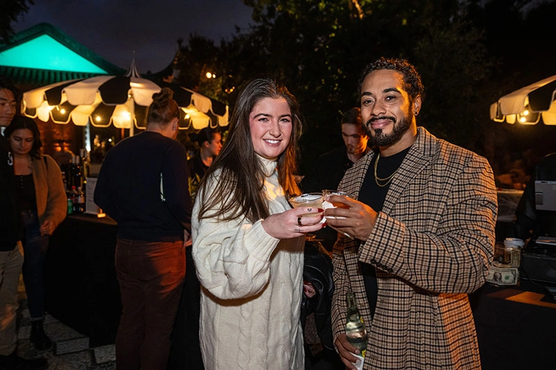 Two people clink drink glasses while posing for the camera.