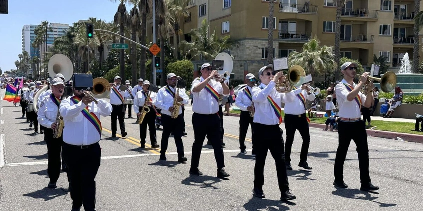 Musicians dressed in black and white with rainbow sashes perform on a sunny day.