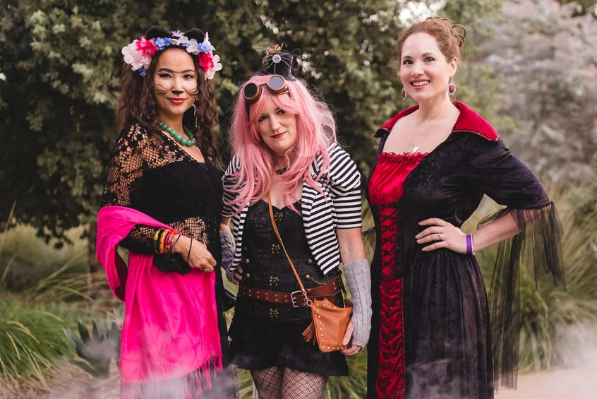 A group of three people stand in a foggy garden wearing black, red, and pink costumes.