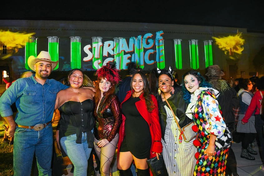 A group of six people in costumes stand in front of a building with a projection that reads "Strange Science."