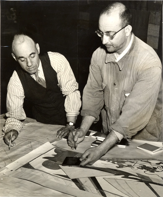 Black and white photo of two men looking down at designs on a table.