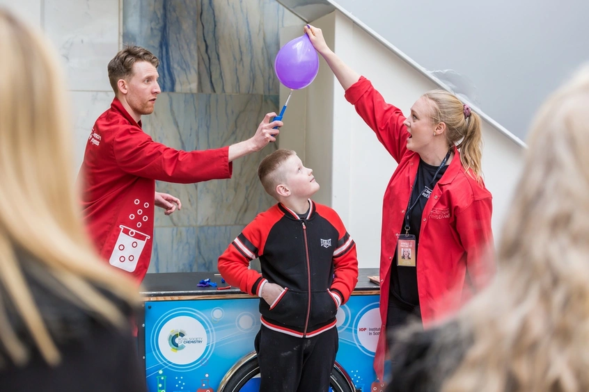 Two people in red lab coats do a balloon experiment over a child.