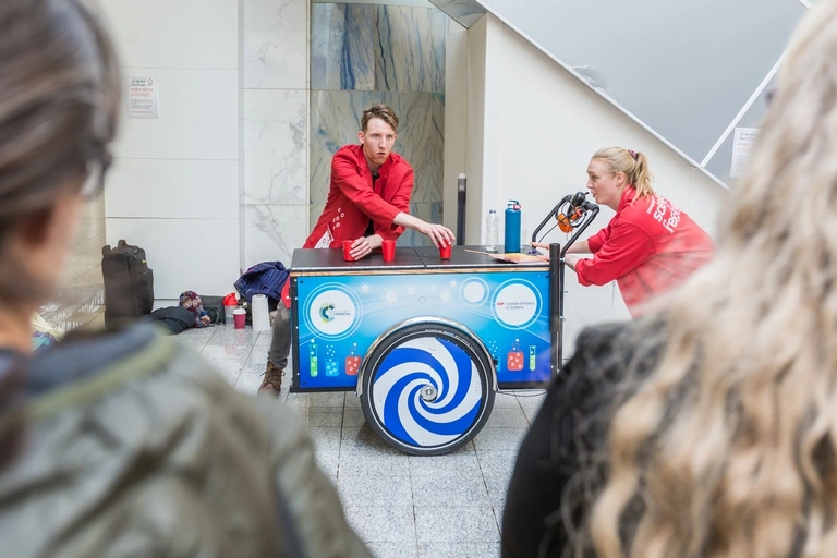 Two people in red coats running a science experiment in front of a crowd.