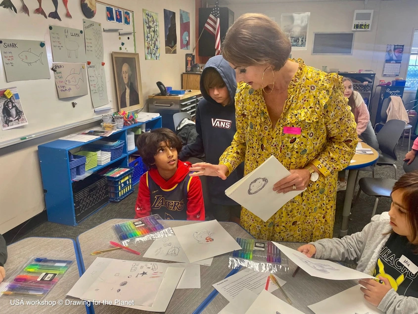 An adult in a yellow dress stands next to a child seated at a table filled with markers and drawings on paper in a busy classroom.