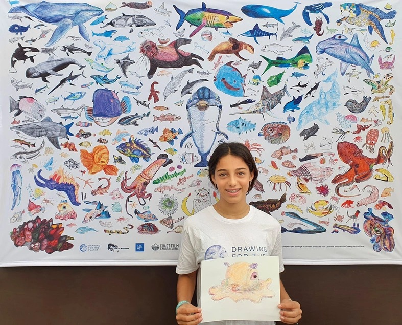 A young person holds a drawing on paper while standing in front of a poster filled with colorful illustrations of sea creatures.