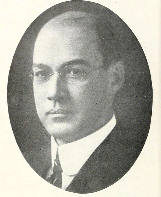 An oval-shaped, faded black-and-white picture of a spectacled man in a suit and tie.