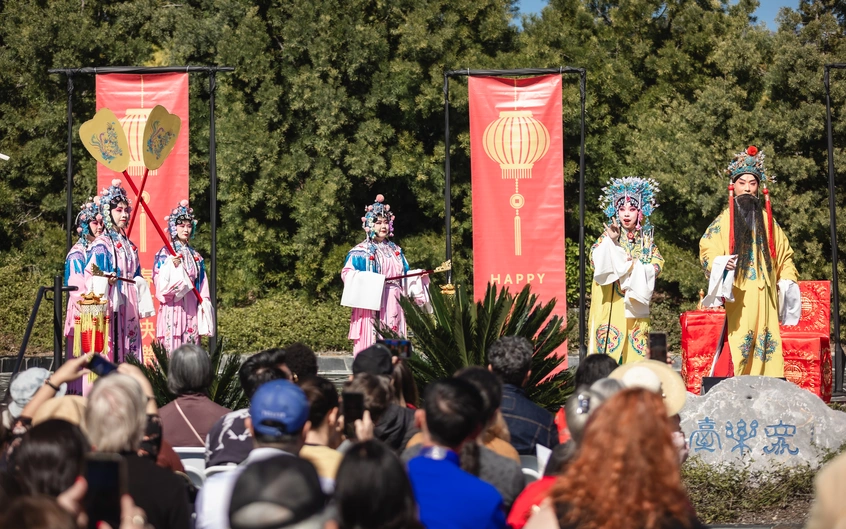 An audience watches six performers wearing intricate, colorful costumes on an outdoor stage.