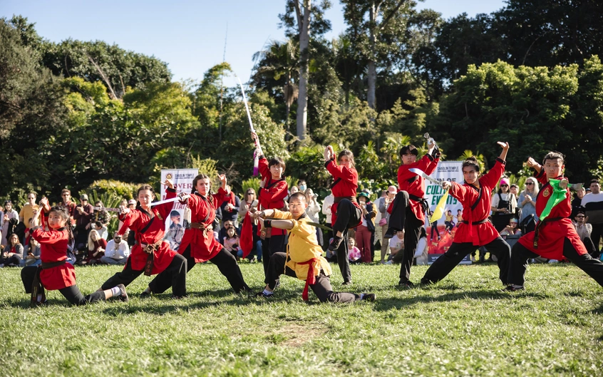 A group of adults in red outfits perform martial arts in front of a crowd on a grass lawn.