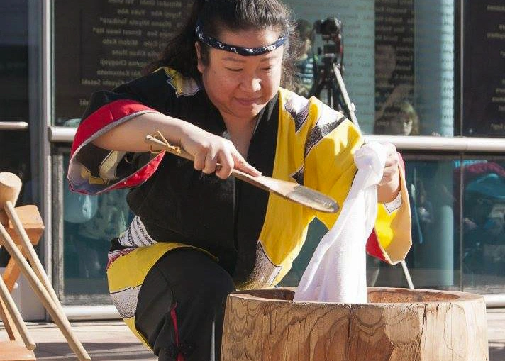 A person squats near a wood bowl, pulling a white substance with their hands and a spoon.