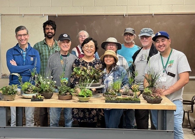 A group of ten people stand behind a table filled with bonsai.