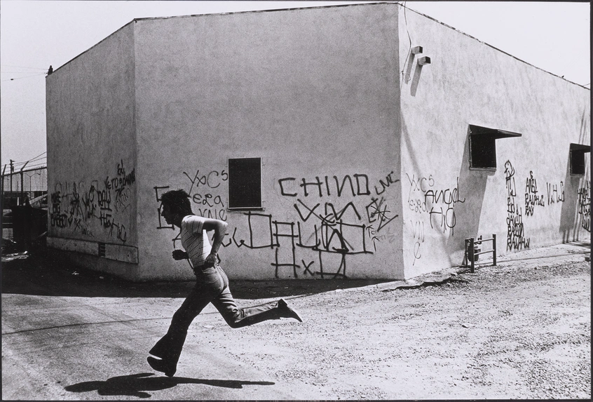 A grayscale image of a person running in front of a gray building with graffiti along the bottom half.