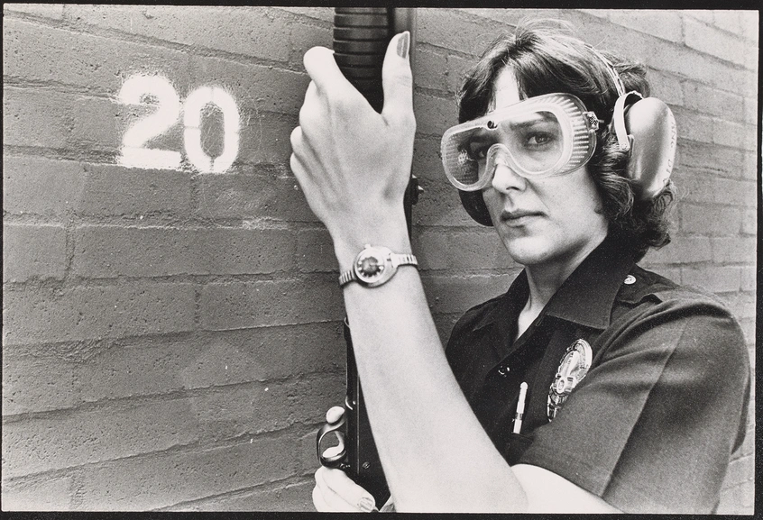 A grayscale image of A person with short hair, wearing safety goggles and earmuffs, holding a gun upright.