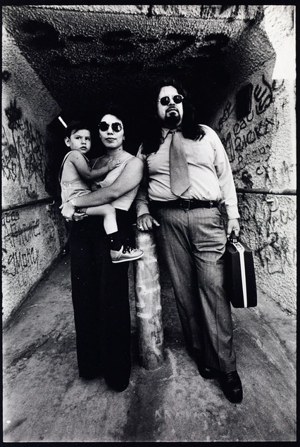 A grayscale image of three people (a man with long hair, sunglasses, and business attire, a woman in a tank top and sunglasses holding a child) standing in front of a dark tunnel filled with graffiti.