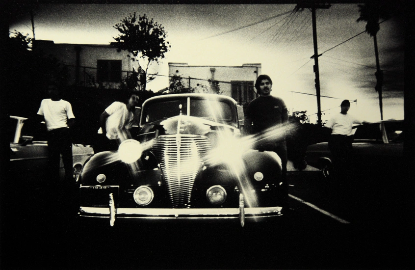 A grayscale image of a 1940's-style muscle car with shining headlights and four people standing nearby.