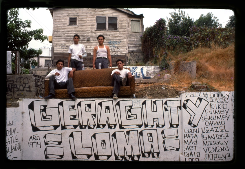 Four people sit on a couch on top of a wall, in a dry landscape, in front of a gray house; the house and wall are covered in graffiti, the largest tag reads "GERAGHTY =LOMA="