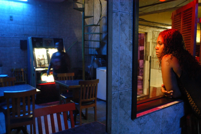 A person with long wavy hair is lit in red light, while leaning into an open window looking into a dining room, with wood chairs and tables, lit in blue light.