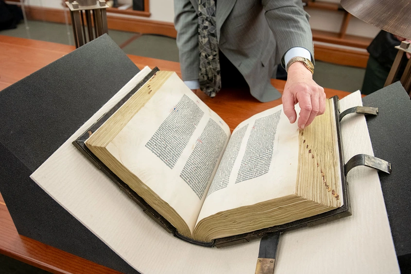 A hand is about to turn the page on an old book sitting open on a book cradle.