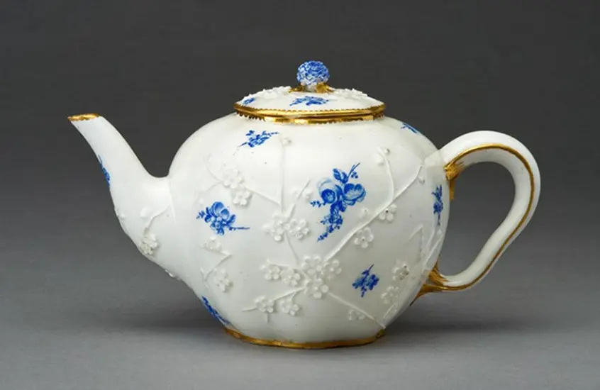 A photo of a teapot from 1740. Pearl white with small, etched flower buds. Blue flower buds highlight the sides and top.