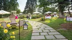 A walkway gently curves to the left through a rose-filled lawn filled with small tables, chairs, and umbrellas.
