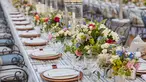 A long dining table filled with flower centerpieces and fancy tableware seems to go on forever as it blurs past the edge of the frame.